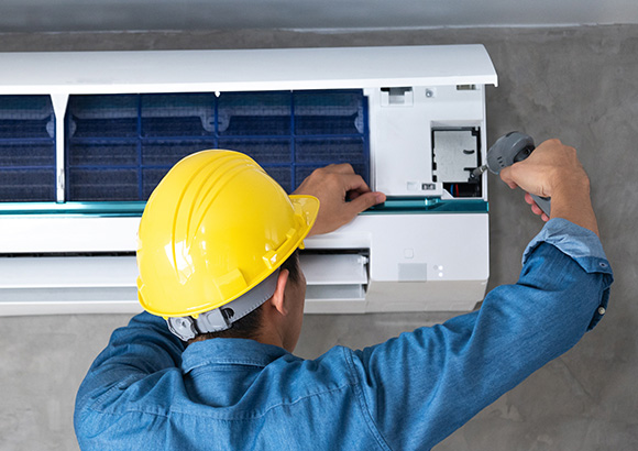 a man wearing a yellow hardhat using an electric screwdriver to perform maintenance on a wall mounted air conditioning unit